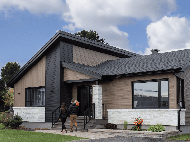 Maison préfabriquée plain-pied du modèle Tryka de style contemporain. Vue de l'extérieur avant. Un couple se dirige vers l'entrée.